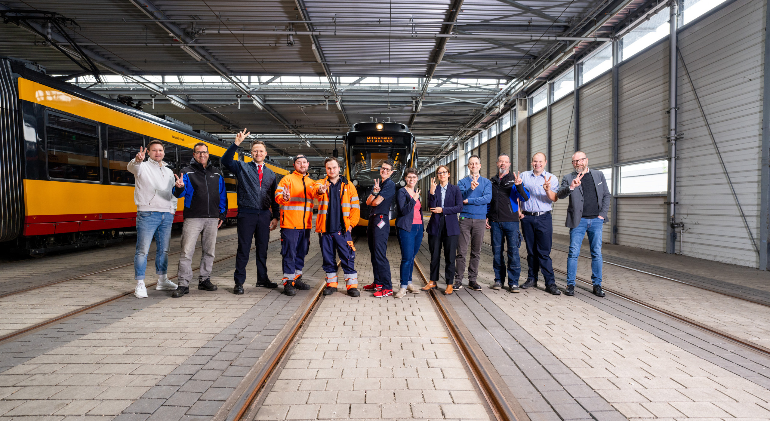 Es stehen zwölf Mitarbeitende, aus unterschiedlichen Bereichen der Unternehmen, nebeneinander mit der #TeamV Pose 