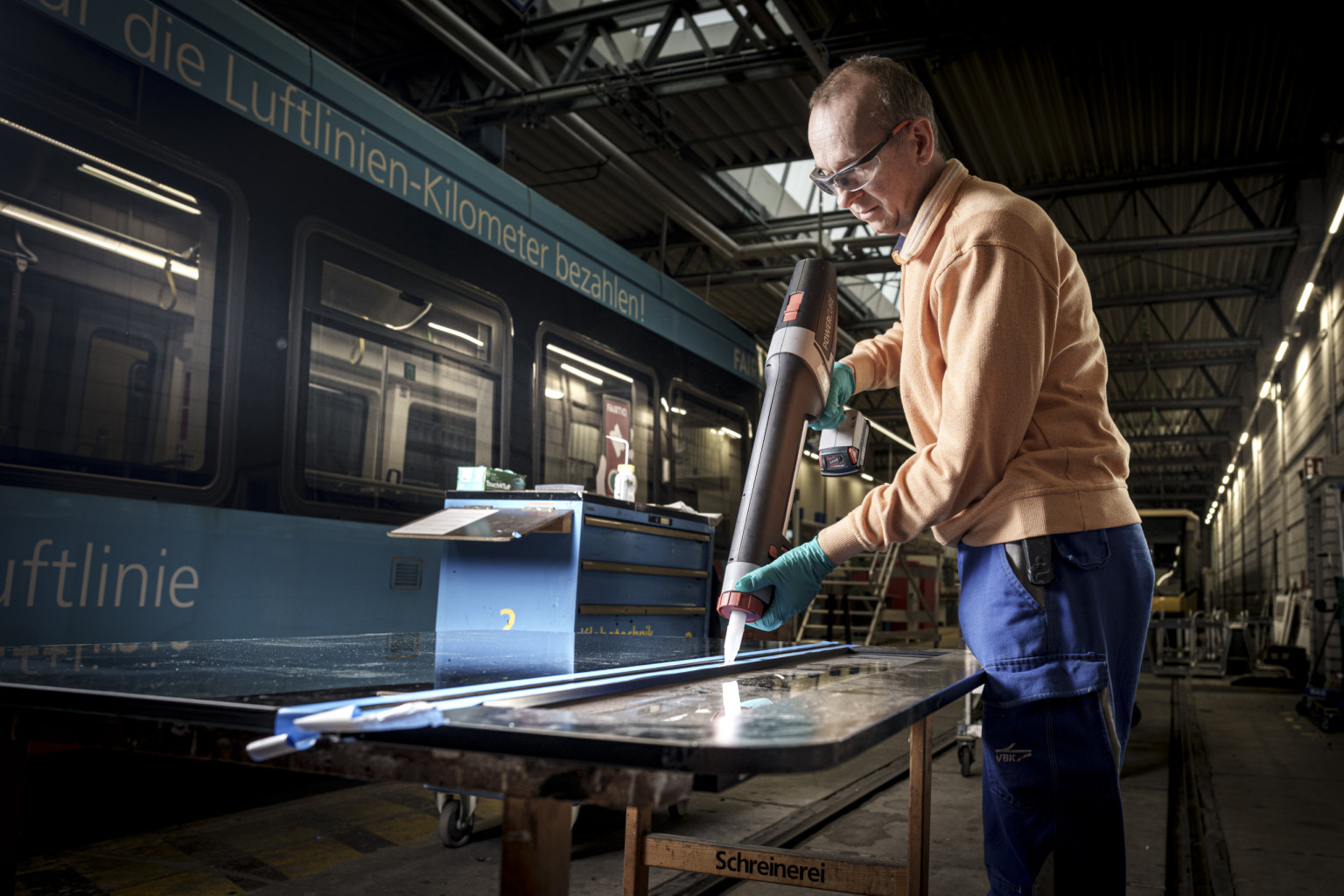 Ein Mitarbeiter aus der Werkstatt repariert ein Bahnteil. Im Hintergrund befindet sich eine blaue Bahn.