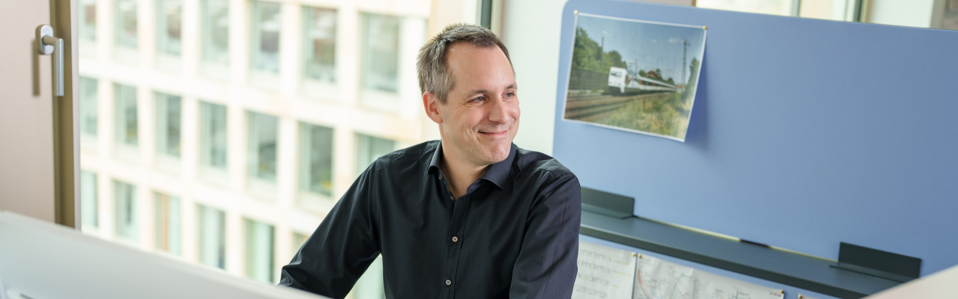 Ein Mitarbeiter steh lächelnd am Arbeitsplatzschreibtisch und schaut nach rechts.